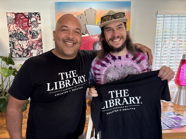 Two smiling attendees display matching black shirts reading “The Library, powered by Kounter Kulture.”