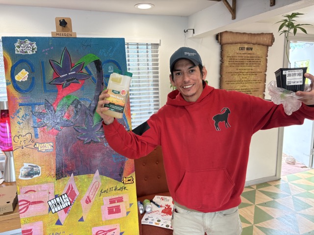 Smiling Clonetopia attendee in a red hoodie holds a bottle of CANNA Start nutrients and a potted cannabis clone.