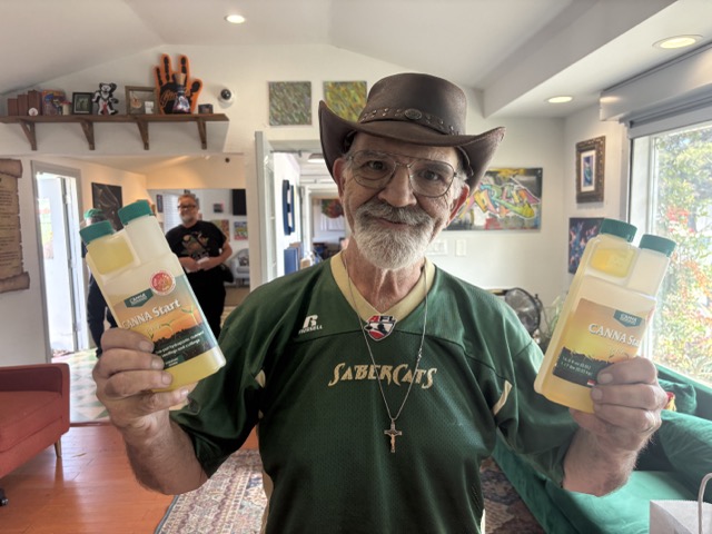 Smiling attendee wearing glasses and a cowboy hat holds two bottles of CANNA Start nutrients at Clonetopia.