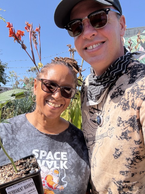 Two smiling Clonetopia attendees pose outdoors, one holding a potted cannabis clone and wearing a Space Walk shirt.