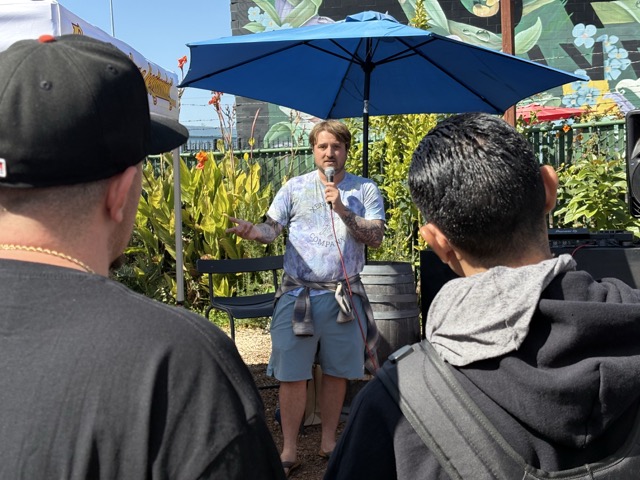 Speaker holds a microphone beneath a blue patio umbrella as Clonetopia attendees listen.