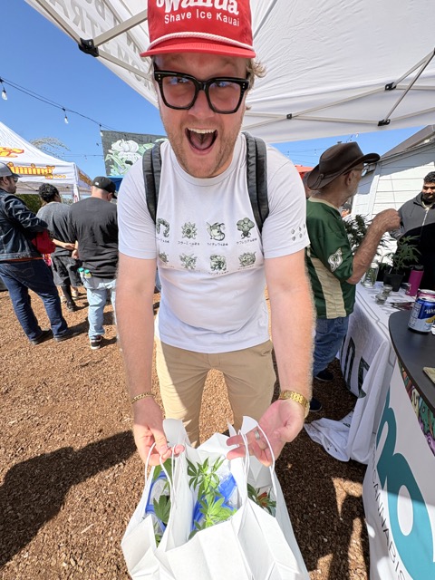 Excited Clonetopia attendee in a red cap holds white shopping bags filled with cannabis clones.